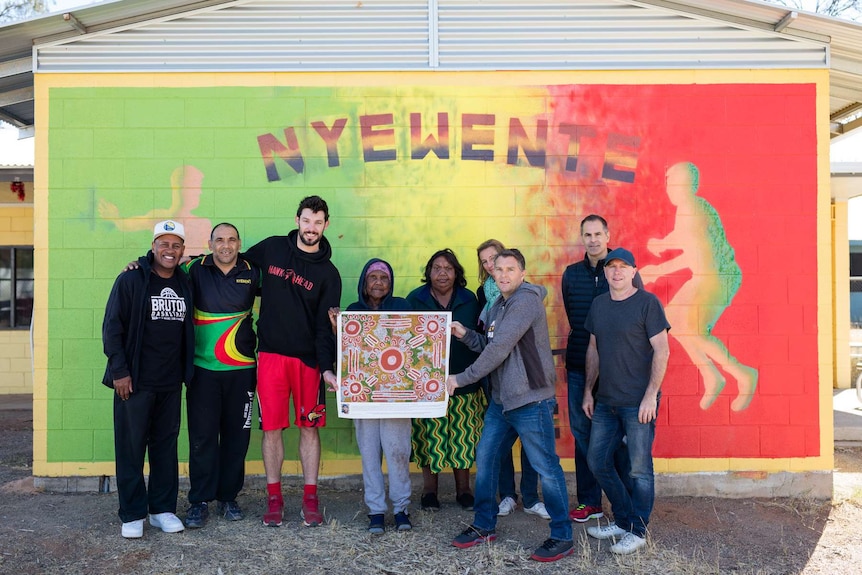 Kevin White poses with Indigenous artwork with community members in front of a mural with Nyewente written on it.
