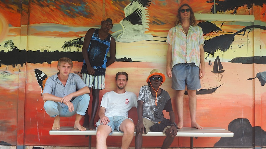 five members of king stingray sit and stand on a bench in front of an orange mural depicting a sunset over water