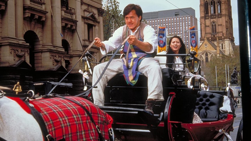 Jackie Chan holds the reins of a horse and carriage looking determined with Miki Lee seated behind, riding through Swanston St.