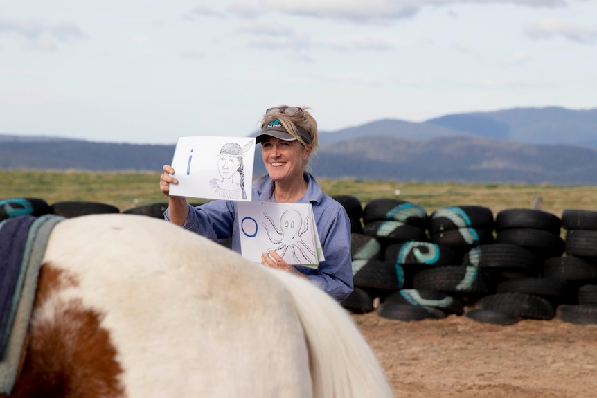 Woman holds up pieces of paper with letters and pictures, while horse stands nearby.