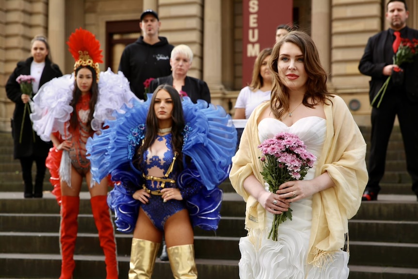 A woman dressed as a bride stands on the steps of a building in front of other people dressed up in colourful costumes.