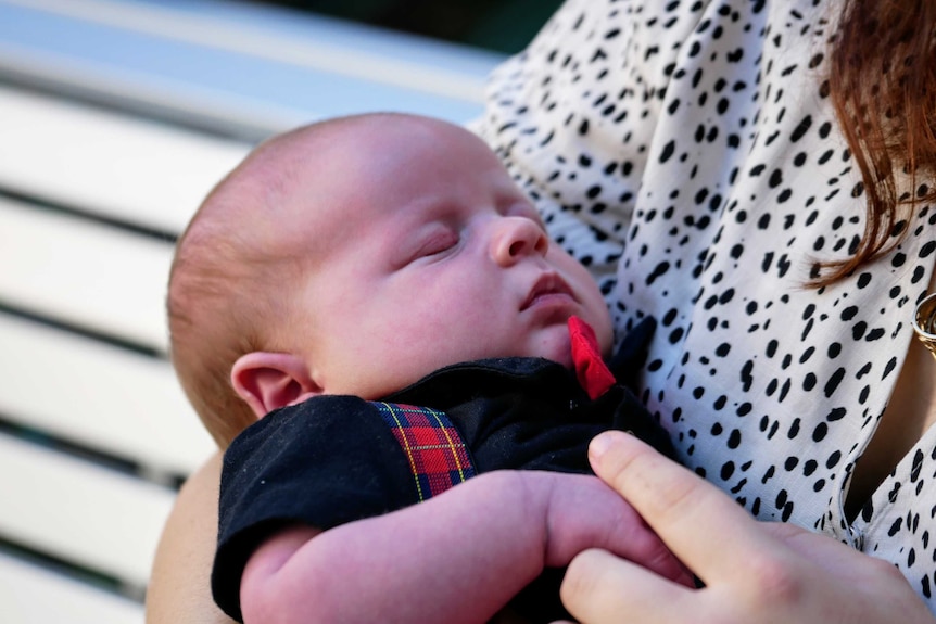 Newborn baby with eyes closed wearing a black shirt, red bowtie, and plaid overalls in mothers arms.