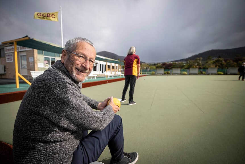 A man in a grey jumper with glasses with a woman on a lawn bowls green in the background.