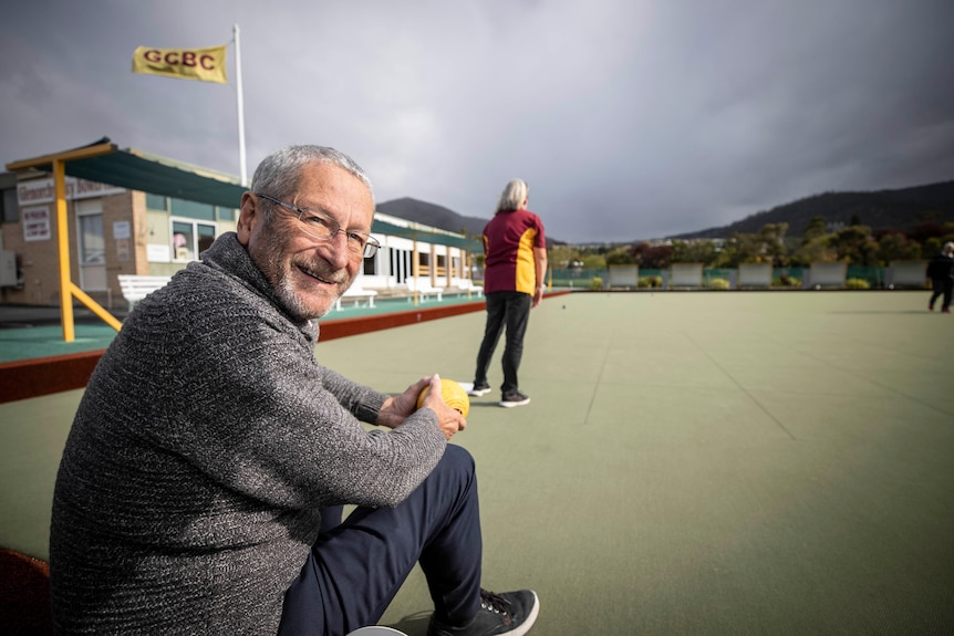 A man in a grey jumper with glasses with a woman on a lawn bowls green in the background.