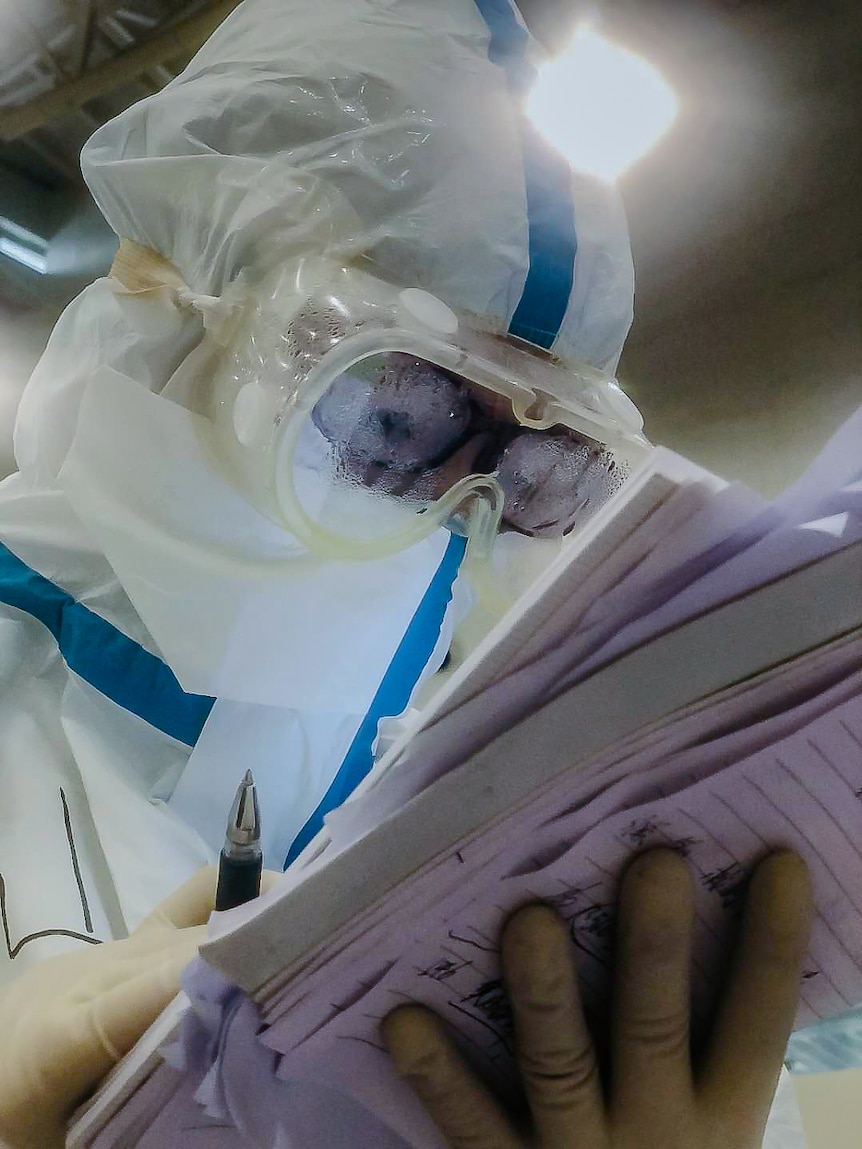 A person in plastic protective gear holding pen and paper with a bright light overhead.