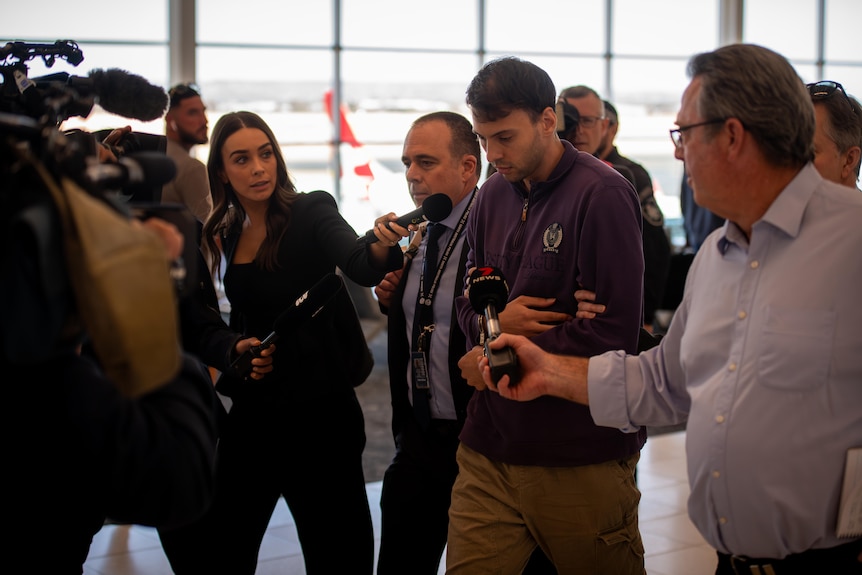 A man in a purple jumper is followed by reporters in an airport.