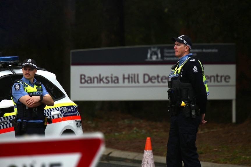 Two police officers stand near a car at the entry to Banksia Hill Juvenile Detention Centre, with a sign marking the entrance.