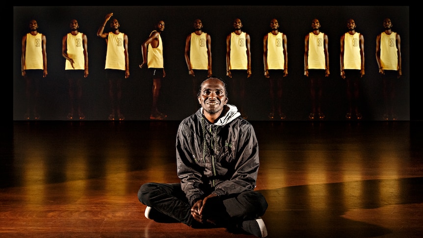 Young man in black pants and hoodie sits on gallery floor with video work featuring ten figures of him standing is behind.