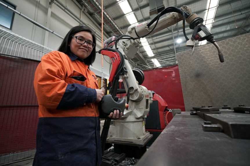 Female manufacturing worker holds a part standing in front of machinery.