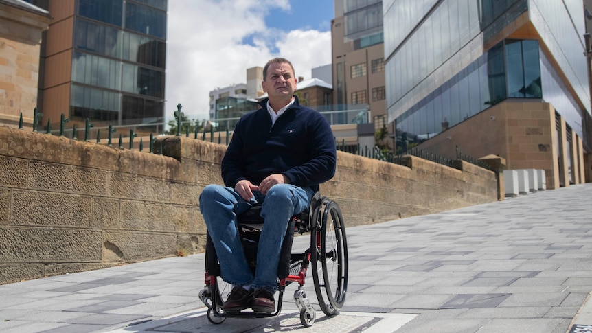 A man sits in a wheelchair on a footpath outside a modern building.