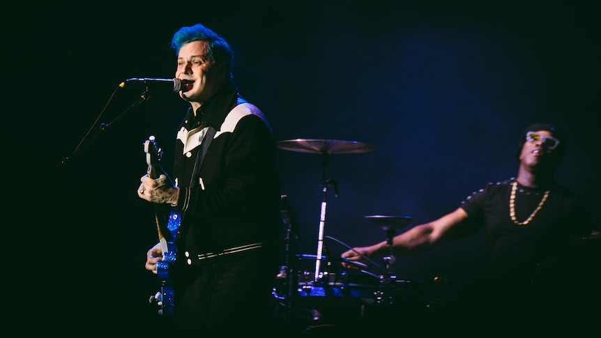Man with short blue hair plays guitar and sings into microphone. Behind him a drummer with a gold chain plays drums
