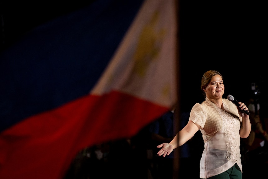 A woman stands in traditional Philippine attire next to the national flag