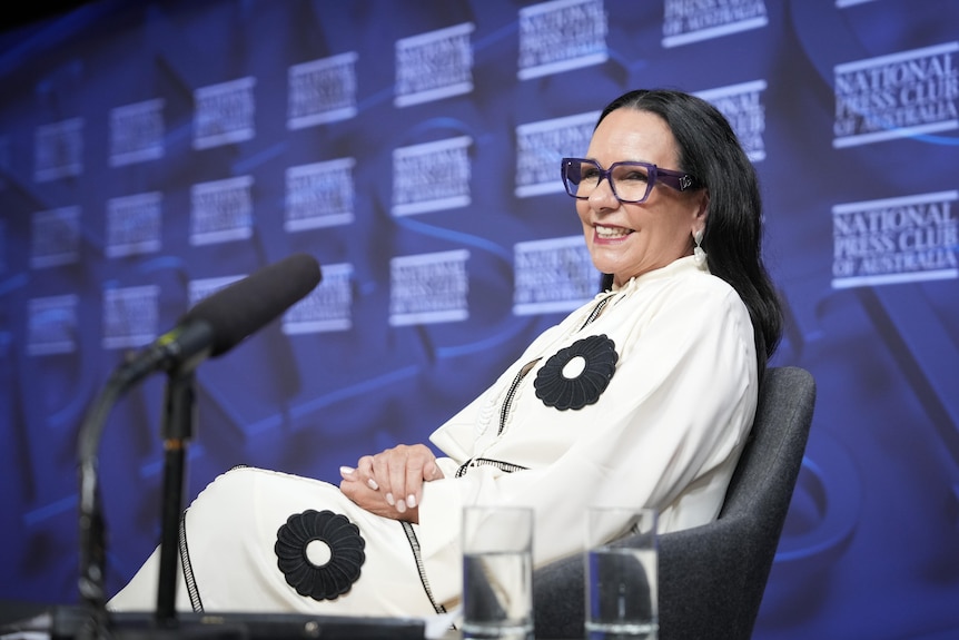 Linda Burney wearing white shirt and purple glasses sitting on a chair and smiling with a blue background