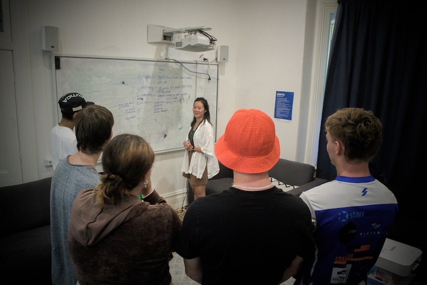 Five young people stand around a woman in front of a whiteboard during a counselling session.
