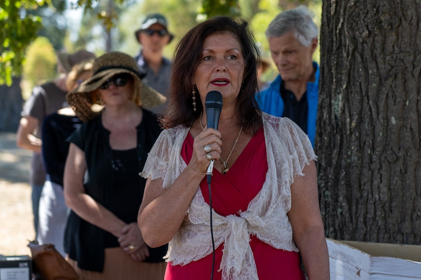 a woman holds a microphone and talks under a tree.