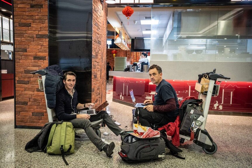 Two men sitting on trolleys in an airport on laptops