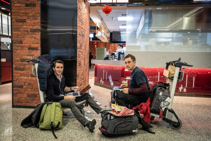 Two men sitting on trolleys in an airport on laptops