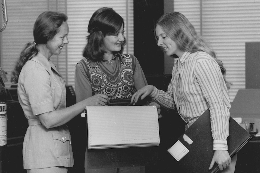 A black and white photo from the 70s showing three women smiling and pointing at a large work book