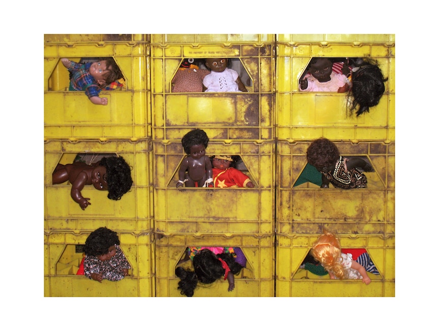 Photographic print by the artist, showing stacked grid of yellow plastic crates filled with small dolls