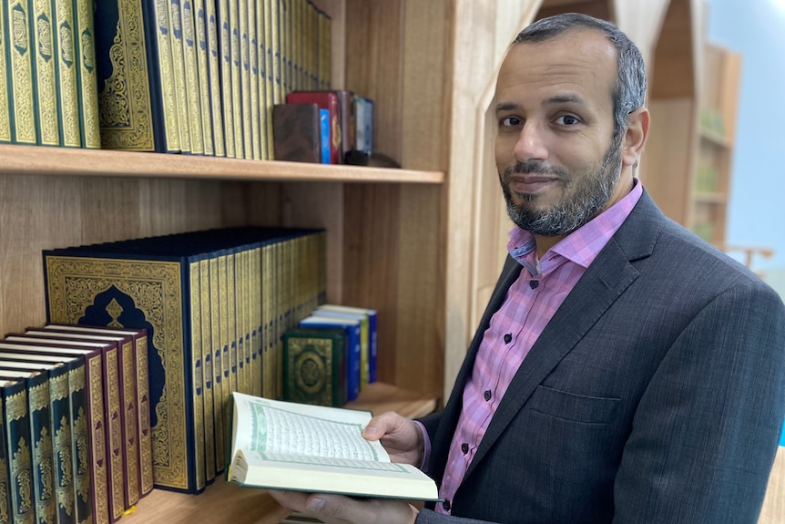 A man stands next to a bookshelf holding an open Koran.