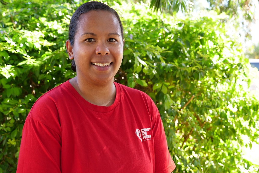 a young woman looking at the camera smiling, wearing a red community spirit foundation shirt, green bush behind.