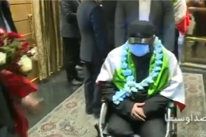 A screenshot of a video shows a man in a wheelchair being surrounded by officials and photographers in dark suits.
