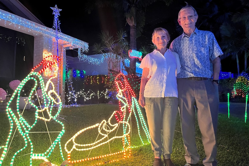 A smiling older Caucasian man and woman standing in front of home decorated with Christmas lights, wears light trousers, shirt.