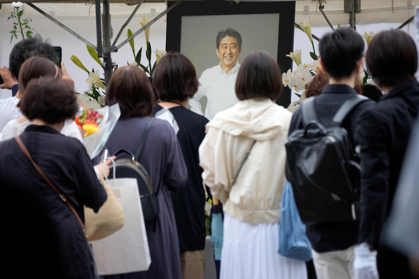 The backs of heads of people are visible looking at a portrait of the late Shinzo Abe smiling.