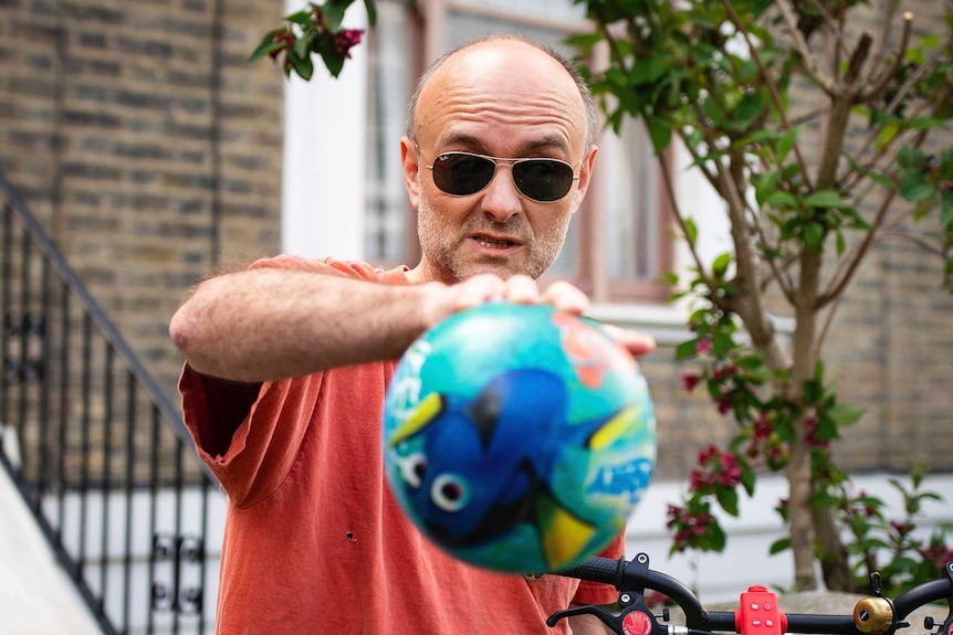 Unshaven , wearing a red t-shirt and sunglass, Dominic Cummings with a Finding Nemo balloon tied to a bicycle.