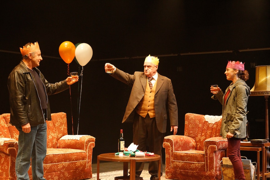 A middle-aged man, an older man and a young woman stand in a living room wearing party hats and cheers their glasses.