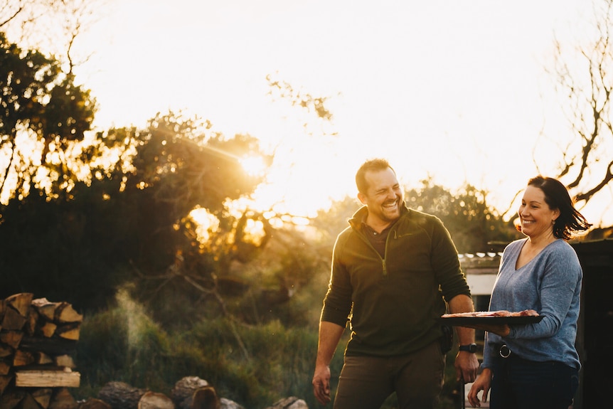 Smiling man and woman walk through setting sun holding plate of steak