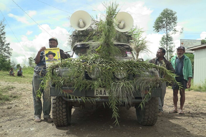Party supporters stand beside a four-wheel drive decorated in green foliage, with two large loudspeakers on top.