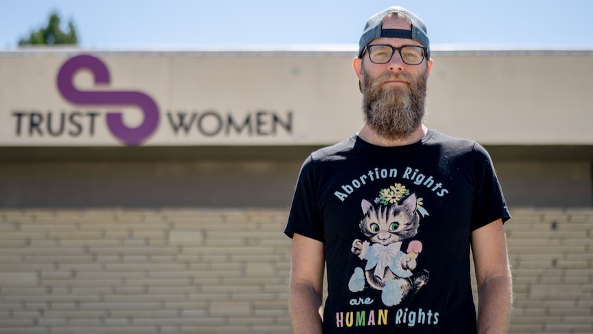 A beareded man wearing a backwards cap and Tshirt that says 'Abortion rights are human rights' stands near a 'Trust Women' sign