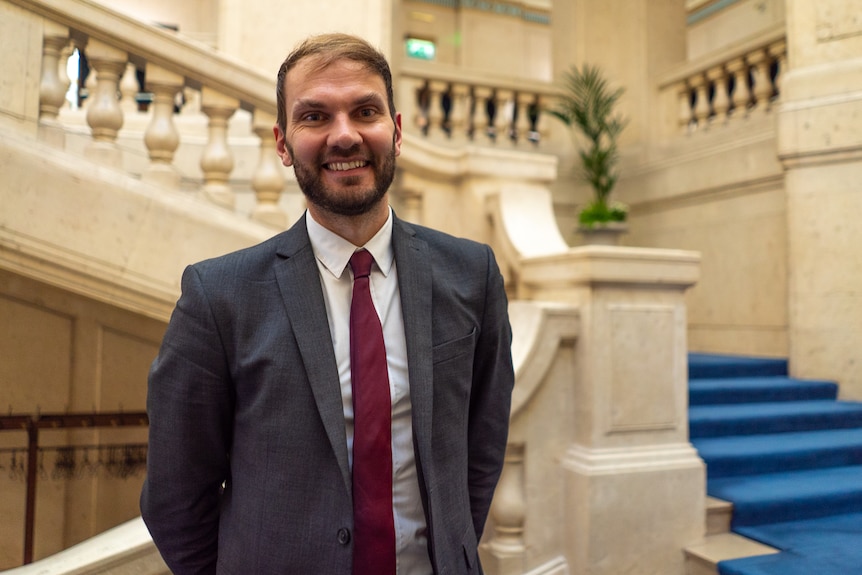 A man with a beard and wearing a grey suit stands on a stairway in a sprawling lobby.