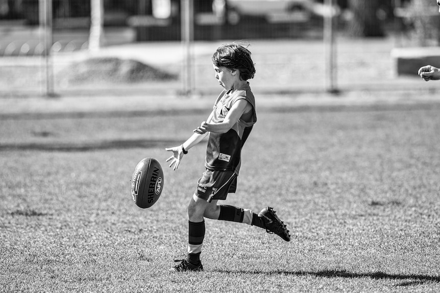 A young boy goes to kick an Aussie Rules ball at a park.