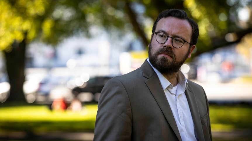 A man with glasses looks to left of image against park-like background.