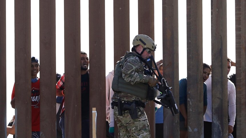 An armed man wearing fatigues and a bulletproof vest walks along one side of a wall while people look on from the other side.