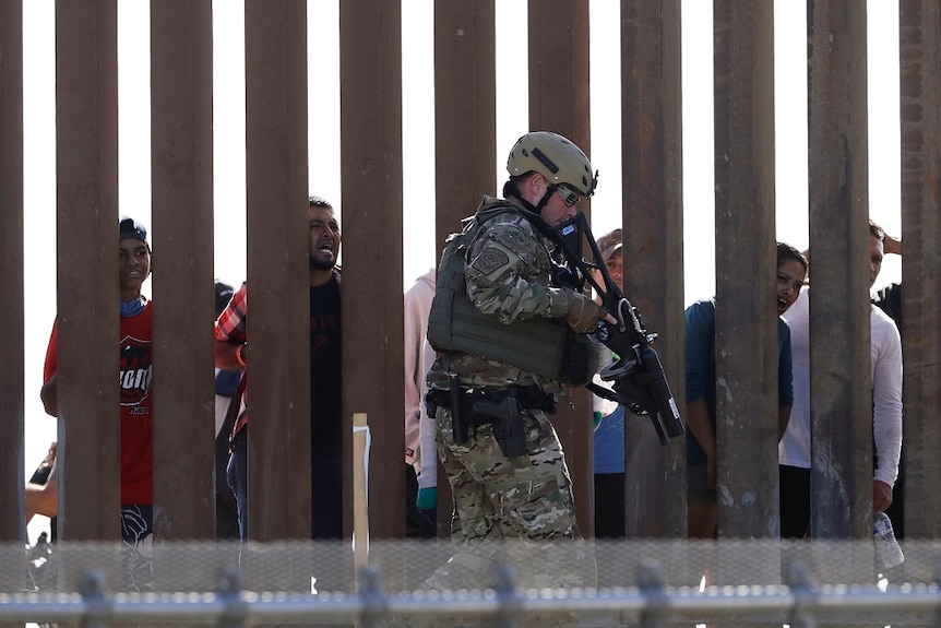 An armed man wearing fatigues and a bulletproof vest walks along one side of a wall while people look on from the other side.