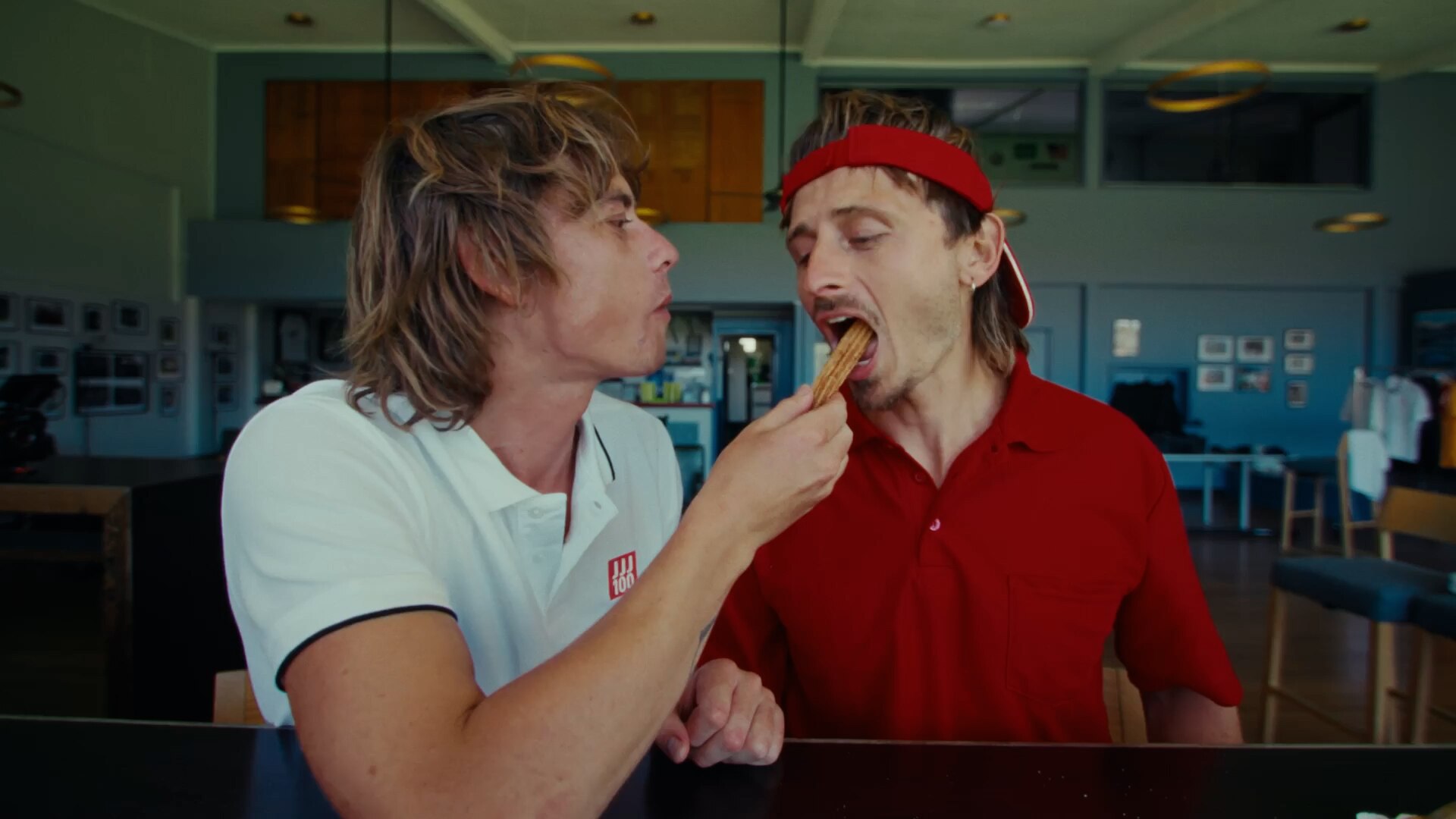 Two men in red and white tennis shirts feed each other churros in a sports clubroom.