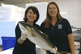 Two smiling women in an industrial-looking area. One holds up a large fish.