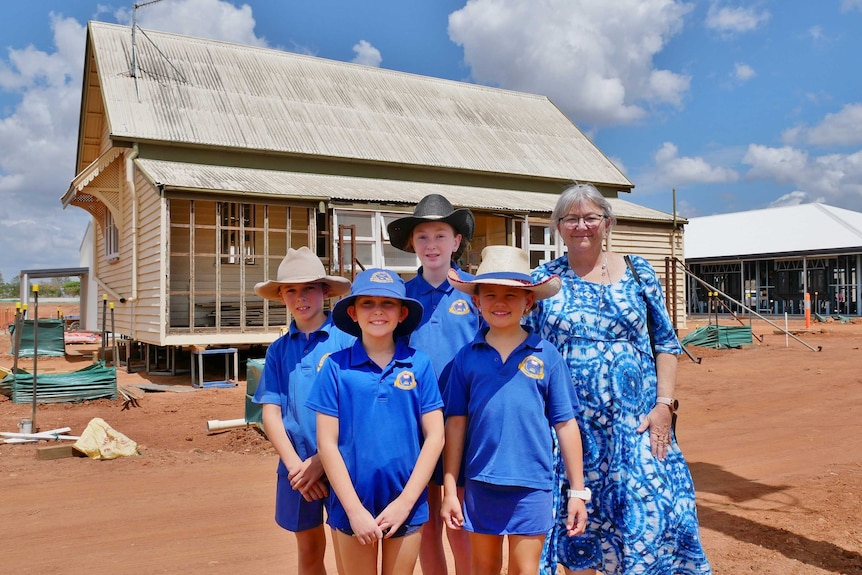 Primary school students in blue uniforms with teacher standing in front of a house in construction.