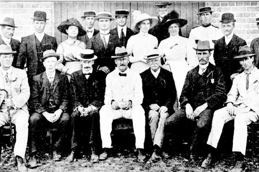 A group of men and women in formal dress sit and stand together, in 1913.