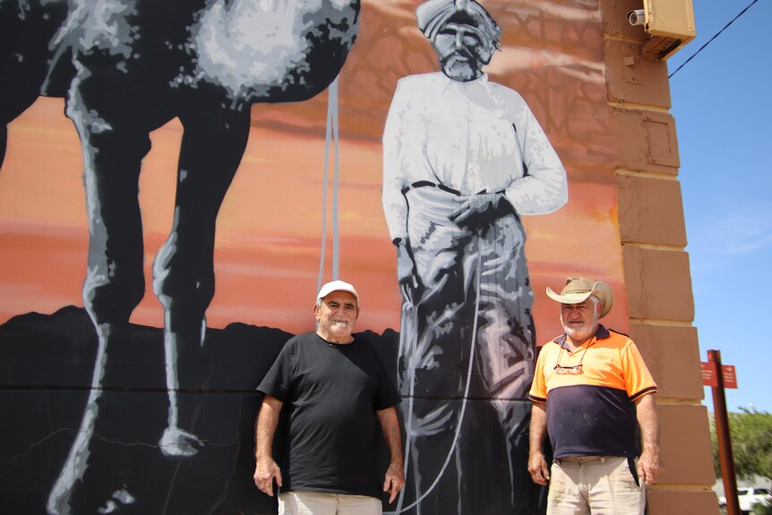 Two men wearing hats standing in front of an art mural depicting a man in a turban and a camel