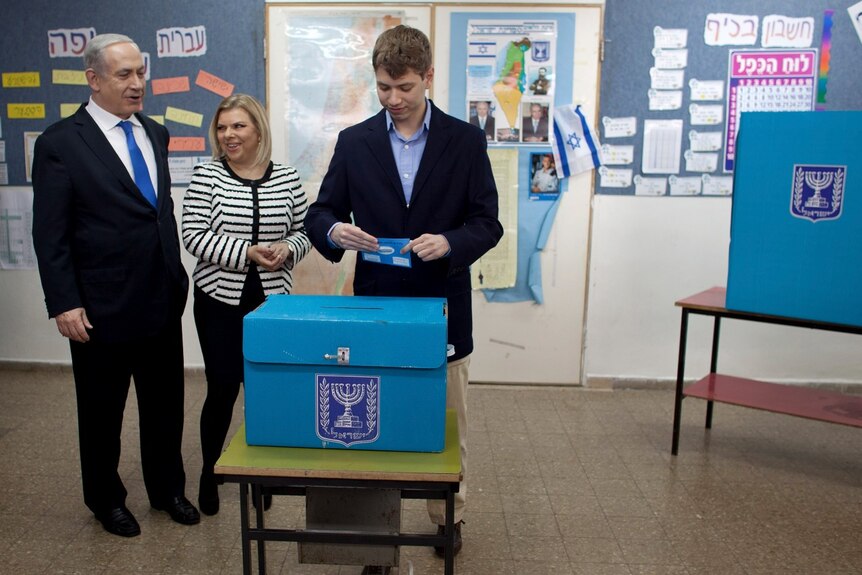 A young man prepares to put a ballot in a box as an older man and woman look on