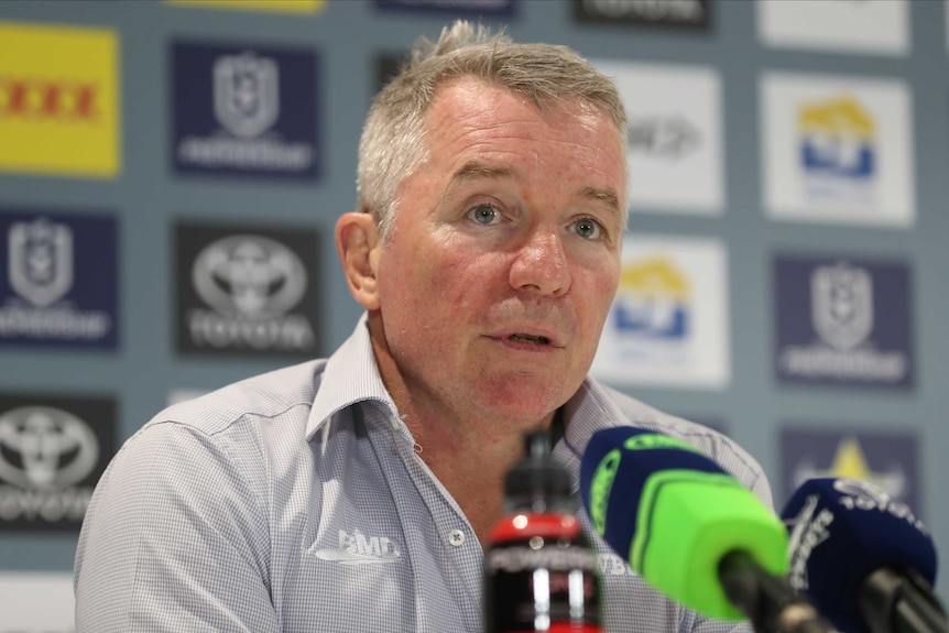 An NRL coach looks out at reporters from behind a desk covered in microphones after a game.