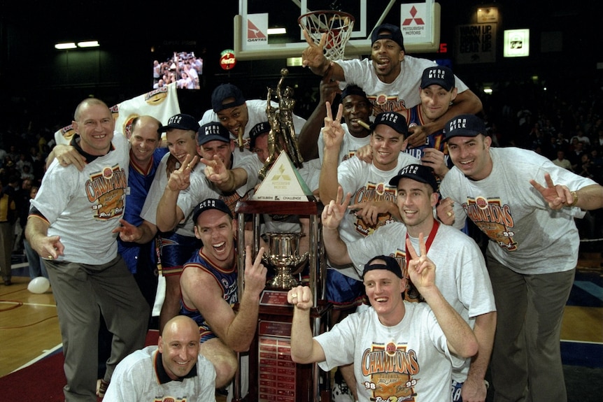 A group of basketball players in Adelaide 36ers gear cheering around a glass-encased trophy on the court