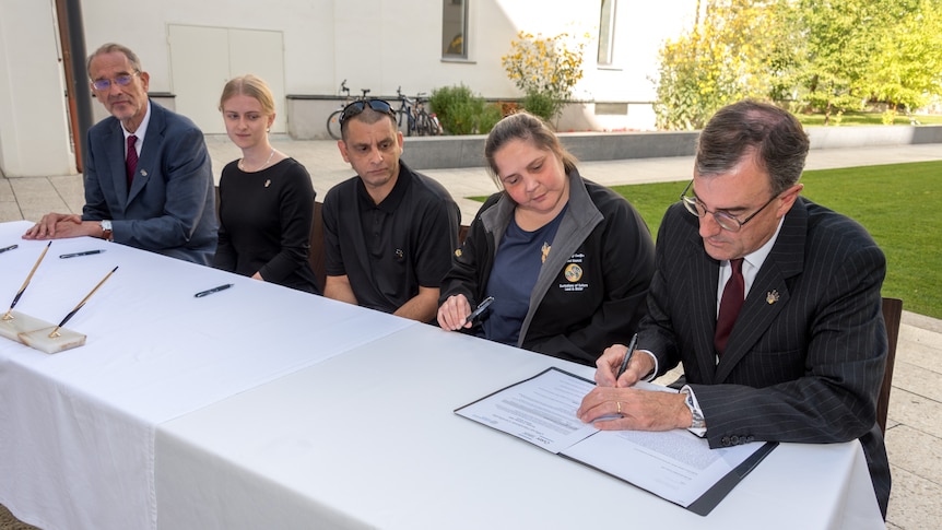a man signs a document, with Indigenous Australians watching on