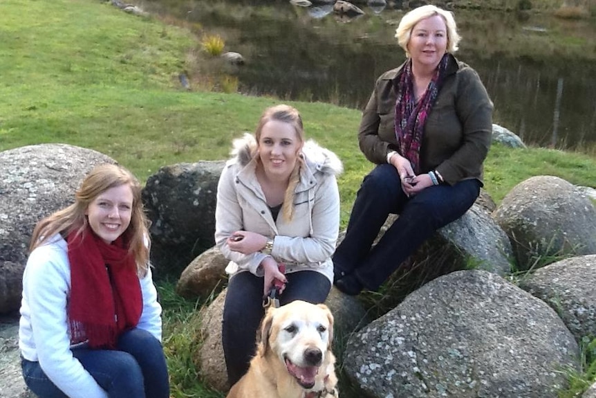 Three women, one holding a dog on leash, sit on rocks, smiling and looking at the camera.