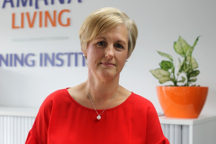 A midshot of a woman wearing a red top, with short blonde hair, stands in front of a white bench and a plant in an orange pot.