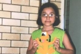 A young girl looks to the  camera with a deadpan expression while wearing a green vest and holding a Scooby Doo toy.